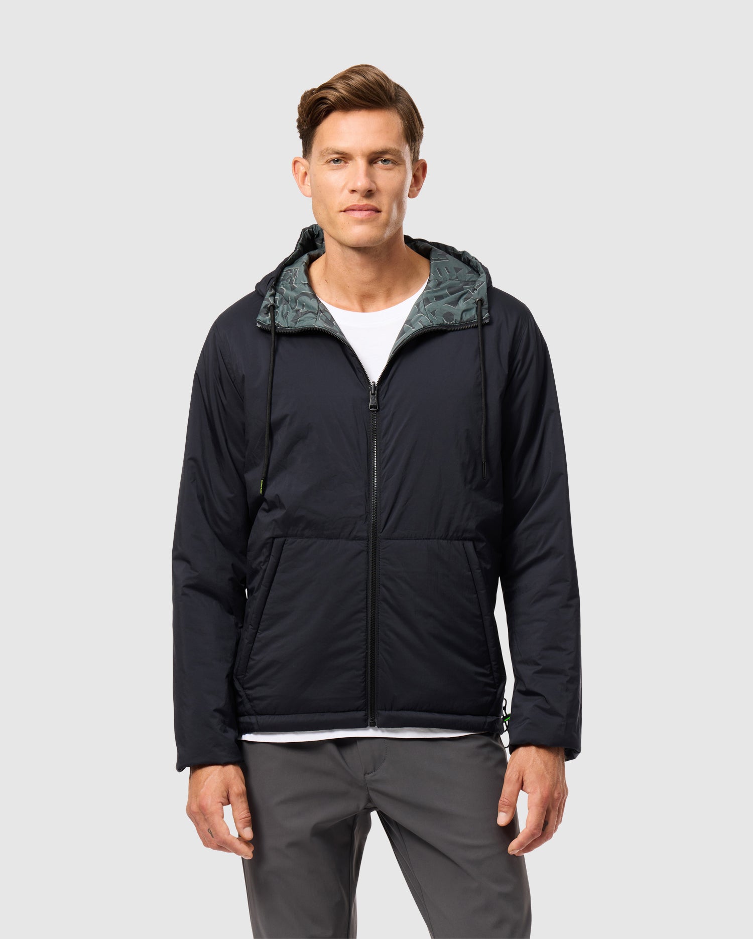 A person wearing the SOUTHWORTH REVERSIBLE PADDED HOODED JACKET (B6J946D200) by Psycho Bunny over a white shirt and gray pants stands against a plain gray background, facing forward with arms relaxed by their sides.