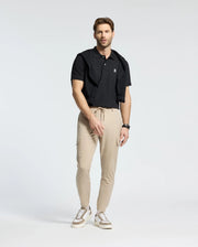 A brown-haired man stands in a studio wearing Psycho Bunny's Men's Classic Pique Polo Shirt in black, with a black sweater draped over his shoulders, beige cargo pants, and white sneakers. He wears a watch on his left wrist and looks forward against a plain white background.