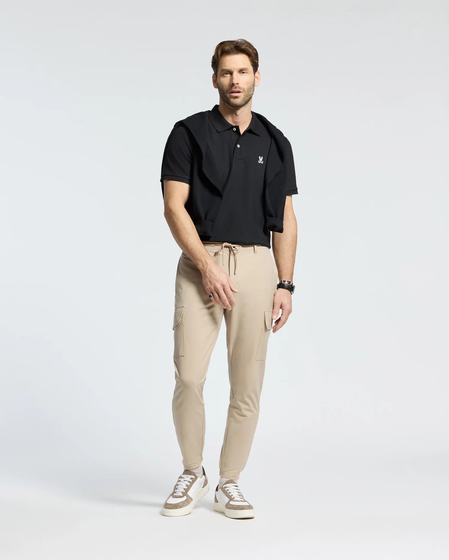 A brown-haired man stands in a studio wearing Psycho Bunny's Men's Classic Pique Polo Shirt in black, with a black sweater draped over his shoulders, beige cargo pants, and white sneakers. He wears a watch on his left wrist and looks forward against a plain white background.