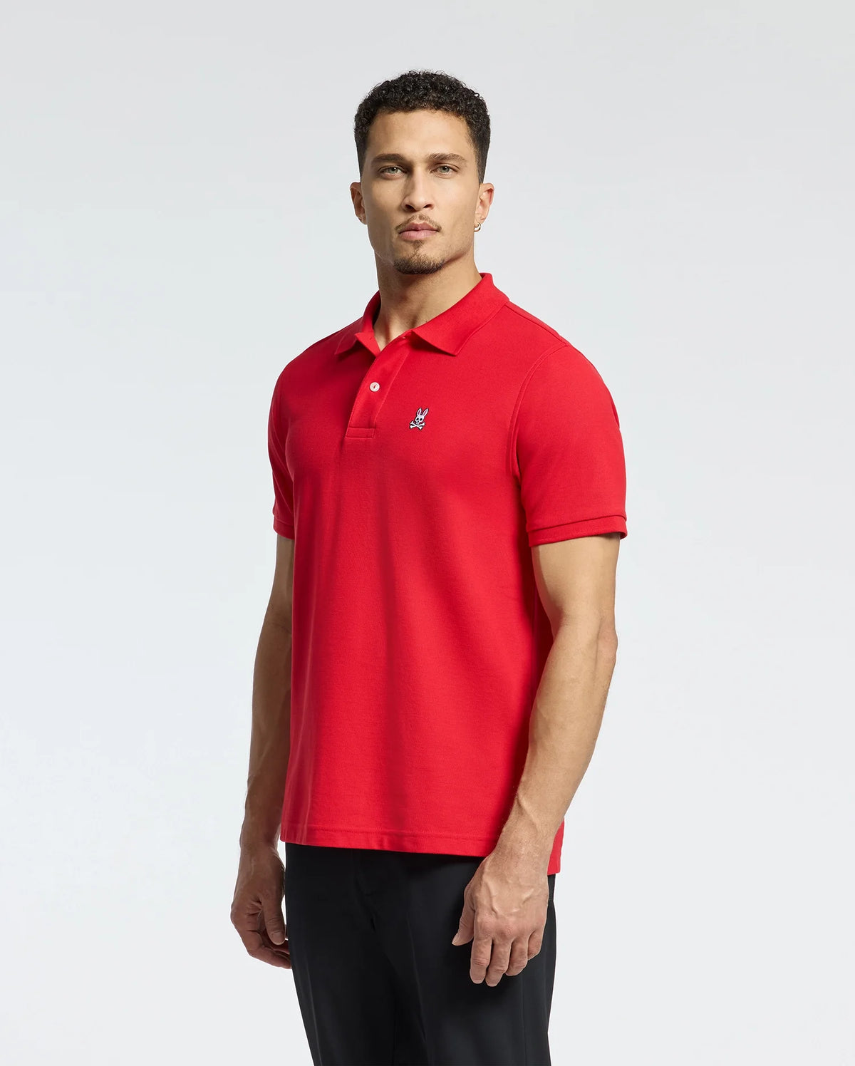 A person is standing against a plain white background, wearing the Psycho Bunny MENS CLASSIC PIQUE POLO SHIRT - B6K001CRPC in red. This polo shirt is made from luxurious Pima cotton with an embroidered bunny logo on the chest and features mother-of-pearl buttons. The individual, who has short dark hair, is gazing directly at the camera and stylishly pairing the polo with black pants.