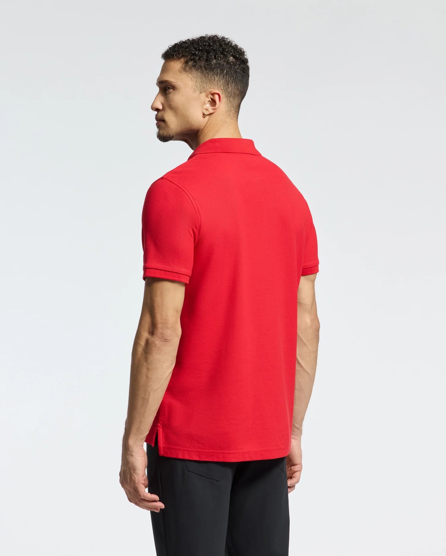 A man with short curly hair is wearing a Psycho Bunny Men's Classic Pique Polo Shirt (B6K001CRPC) in red, paired with black pants. He is standing sideways, facing to the left, against a plain and light-colored background.