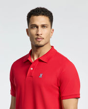A man with short curly hair wears a vibrant red Psycho Bunny Men's Classic Pique Polo Shirt, crafted from soft Pima cotton and adorned with mother-of-pearl buttons and a small embroidered logo. The plain, light-colored background accentuates the vibrancy of his clothing.