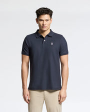 A person stands with short dark hair, wearing a Psycho Bunny Men's Classic Pique Polo Shirt in classic navy, featuring a small embroidered logo on the left chest. Made from Pima cotton for added comfort and style, they're also in light-colored pants against a plain light gray background.