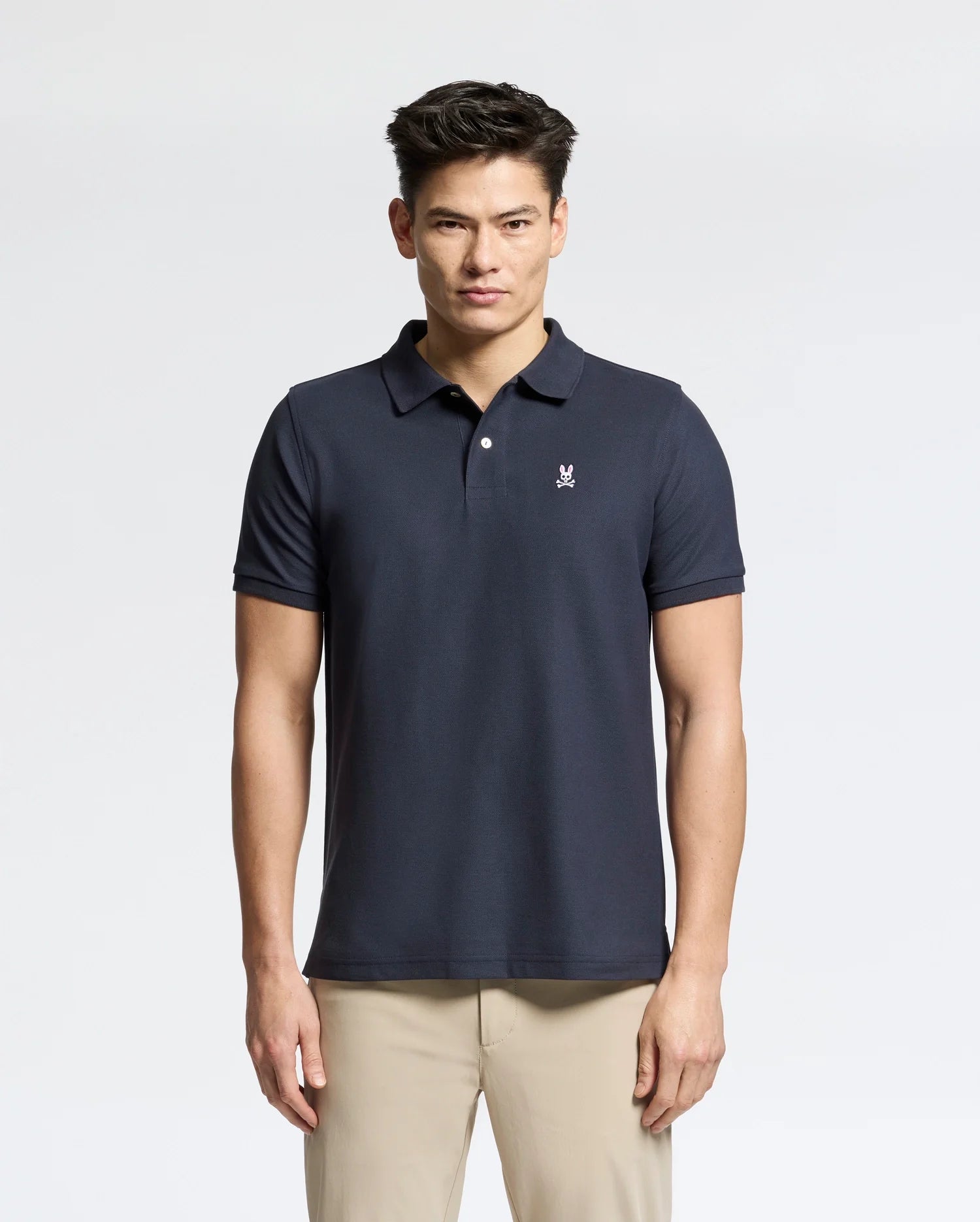 A person stands with short dark hair, wearing a Psycho Bunny Men's Classic Pique Polo Shirt in classic navy, featuring a small embroidered logo on the left chest. Made from Pima cotton for added comfort and style, they're also in light-colored pants against a plain light gray background.