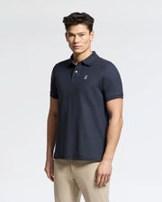 A man with short, dark hair wears a Psycho Bunny Men's Classic Pique Polo Shirt (B6K001ARPC) crafted from Pima cotton, sporting a small embroidered logo on the chest. He pairs it with beige pants against a plain light-colored background.