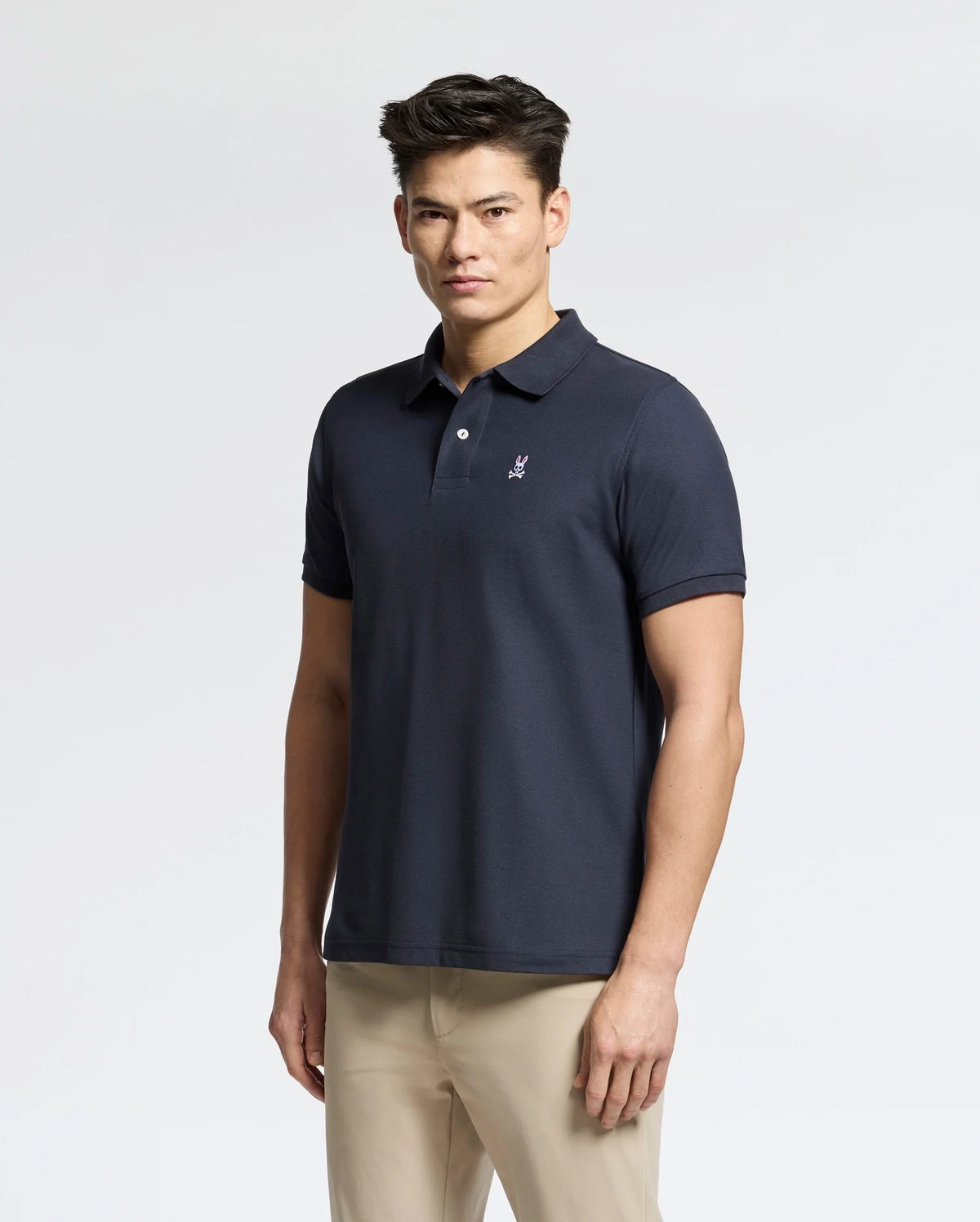 A man with short, dark hair wears a Psycho Bunny Men's Classic Pique Polo Shirt (B6K001ARPC) crafted from Pima cotton, sporting a small embroidered logo on the chest. He pairs it with beige pants against a plain light-colored background.