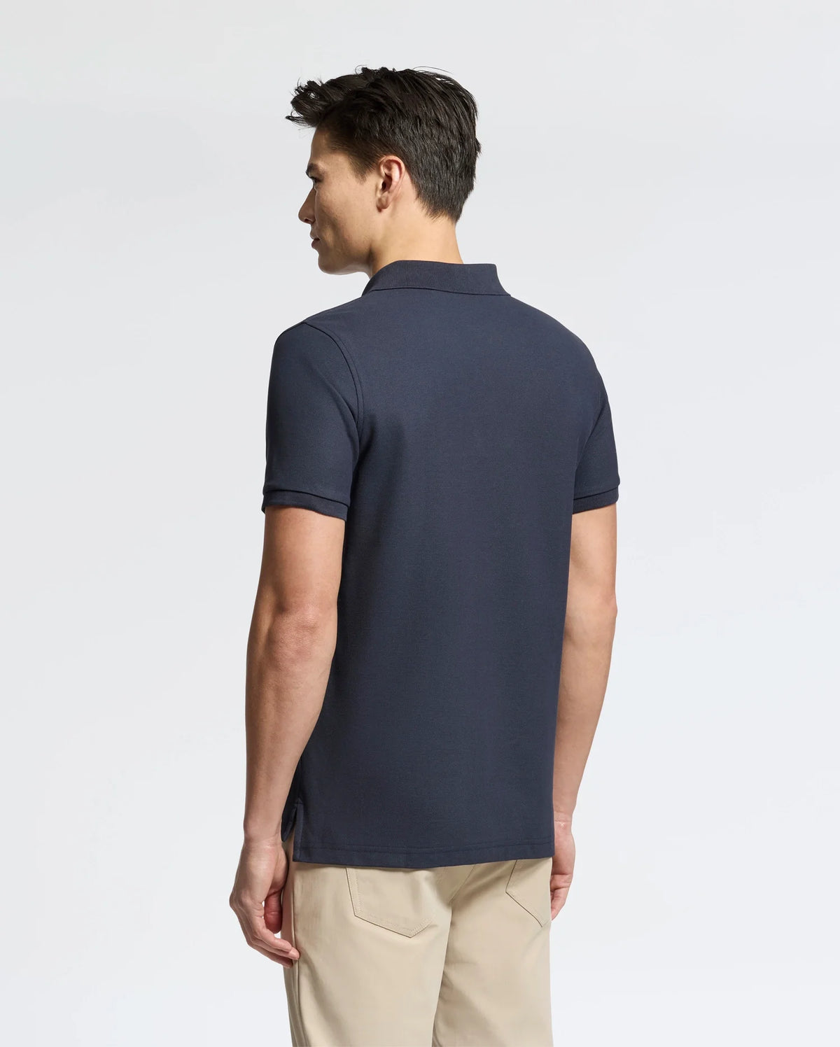 A person with short, dark hair is wearing the Psycho Bunny MENS CLASSIC PIQUE POLO SHIRT - B6K001ARPC in dark grey, paired with light beige pants. They are standing slightly turned to the left against a plain background.