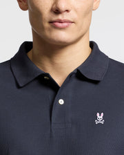 A man wears the Psycho Bunny Men's Classic Pique Polo Shirt - B6K001ARPC, made from soft Pima cotton in a dark color, featuring a pink-eared bunny skull emblem on crossed bones. The buttoned collar stands out against the plain white background.