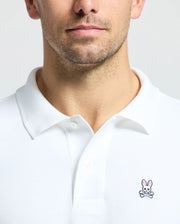 A man is wearing a Psycho Bunny Men's Classic Pique Polo Shirt - B6K001ARPC, made from 100% Pima cotton. It features a subtle embroidered bunny and crossbones logo on the chest and elegant mother-of-pearl buttons. The plain gray backdrop enhances the shirt's sophisticated simplicity.