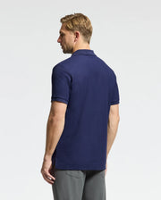 A man with short blond hair stands looking to the side, dressed in a Psycho Bunny navy blue MENS CLASSIC PIQUE POLO SHIRT - B6K001CRPC and gray trousers. The background is plain white.