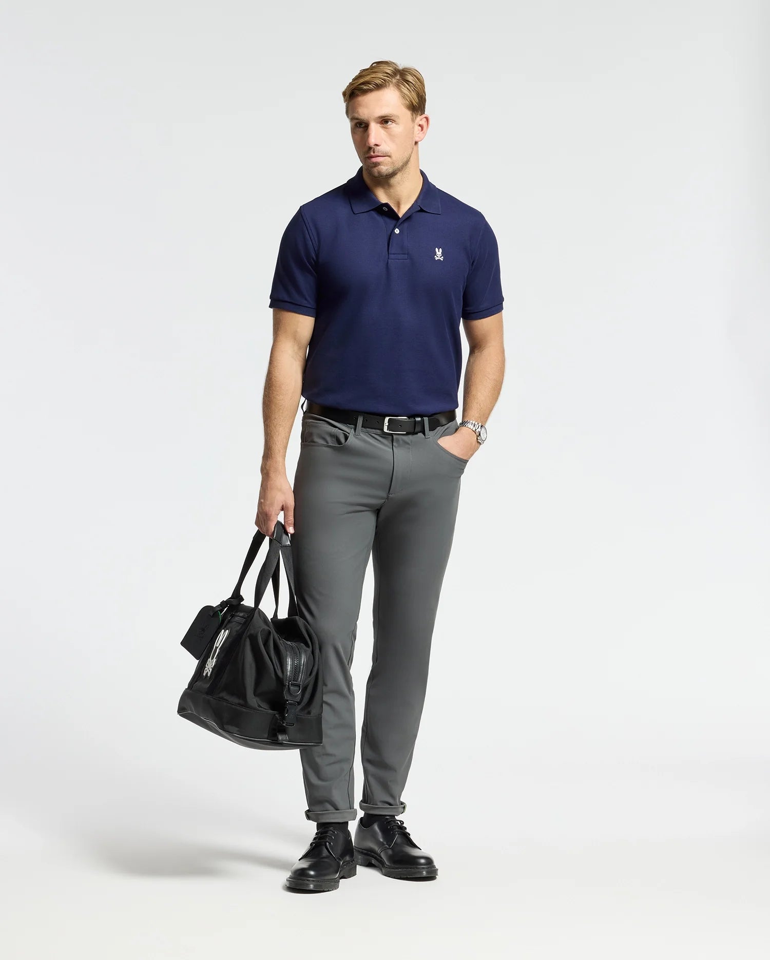 A man wearing a Psycho Bunny MENS CLASSIC PIQUE POLO SHIRT - B6K001CRPC in navy pairs it with gray pants and holds a black duffel bag. He accessorizes with a wristwatch while standing in front of a plain white background.