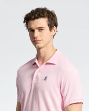 A person with curly hair wears a pale pink Psycho Bunny MENS CLASSIC PIQUE POLO SHIRT - B6K001CRPC, featuring a small embroidered logo on the chest, against a neutral background. The Pima cotton adds comfort as they look toward the camera with a slight smile.