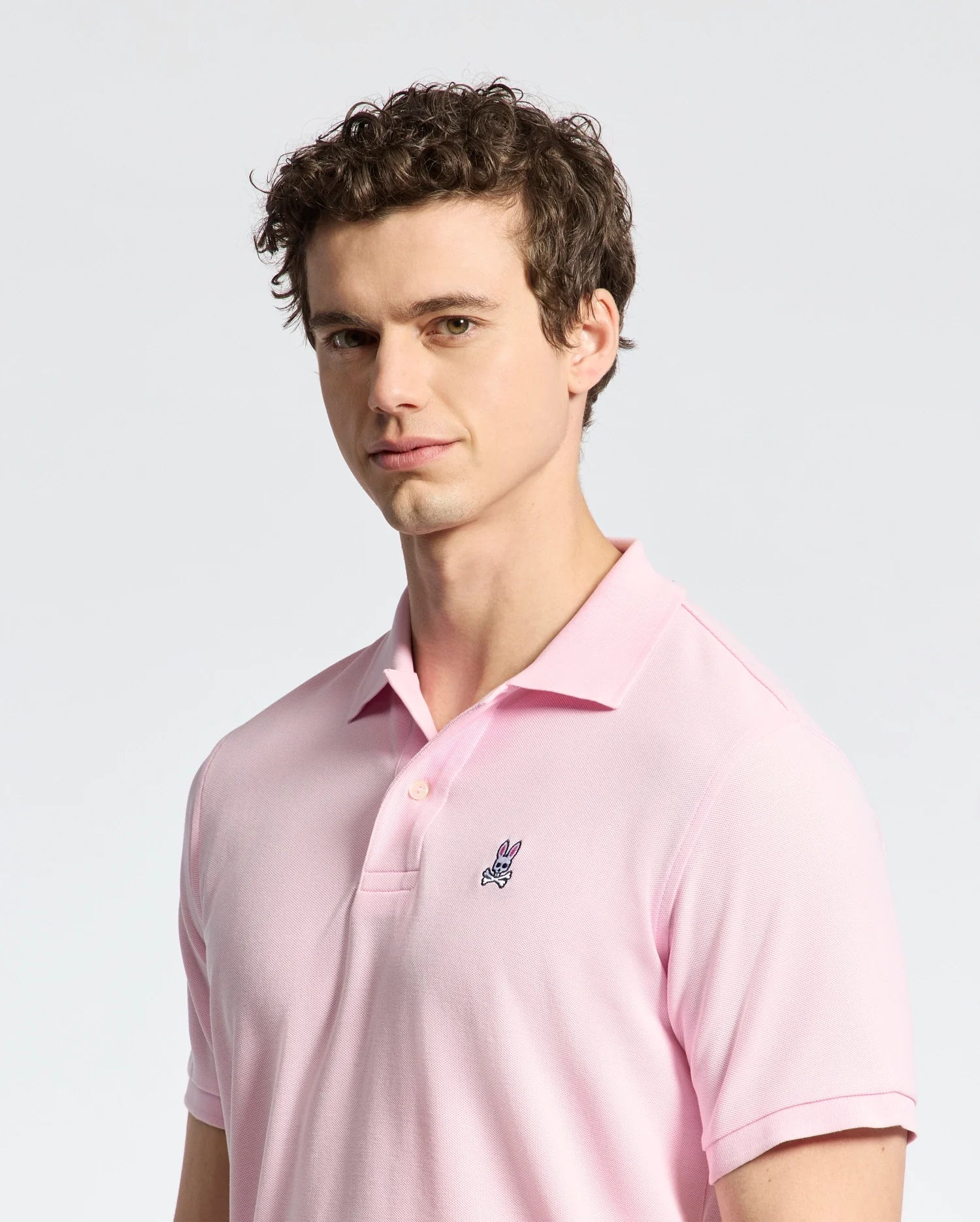 A person with curly hair wears a pale pink Psycho Bunny MENS CLASSIC PIQUE POLO SHIRT - B6K001CRPC, featuring a small embroidered logo on the chest, against a neutral background. The Pima cotton adds comfort as they look toward the camera with a slight smile.
