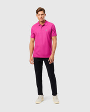 A person wearing the Psycho Bunny MENS CLASSIC PIQUE POLO SHIRT - B6K001D200 in a vibrant pink hue stands against a plain background. Paired with black pants and white sneakers, the 100% Pima cotton fabric exudes subtle elegance as they gaze ahead with a neutral expression.