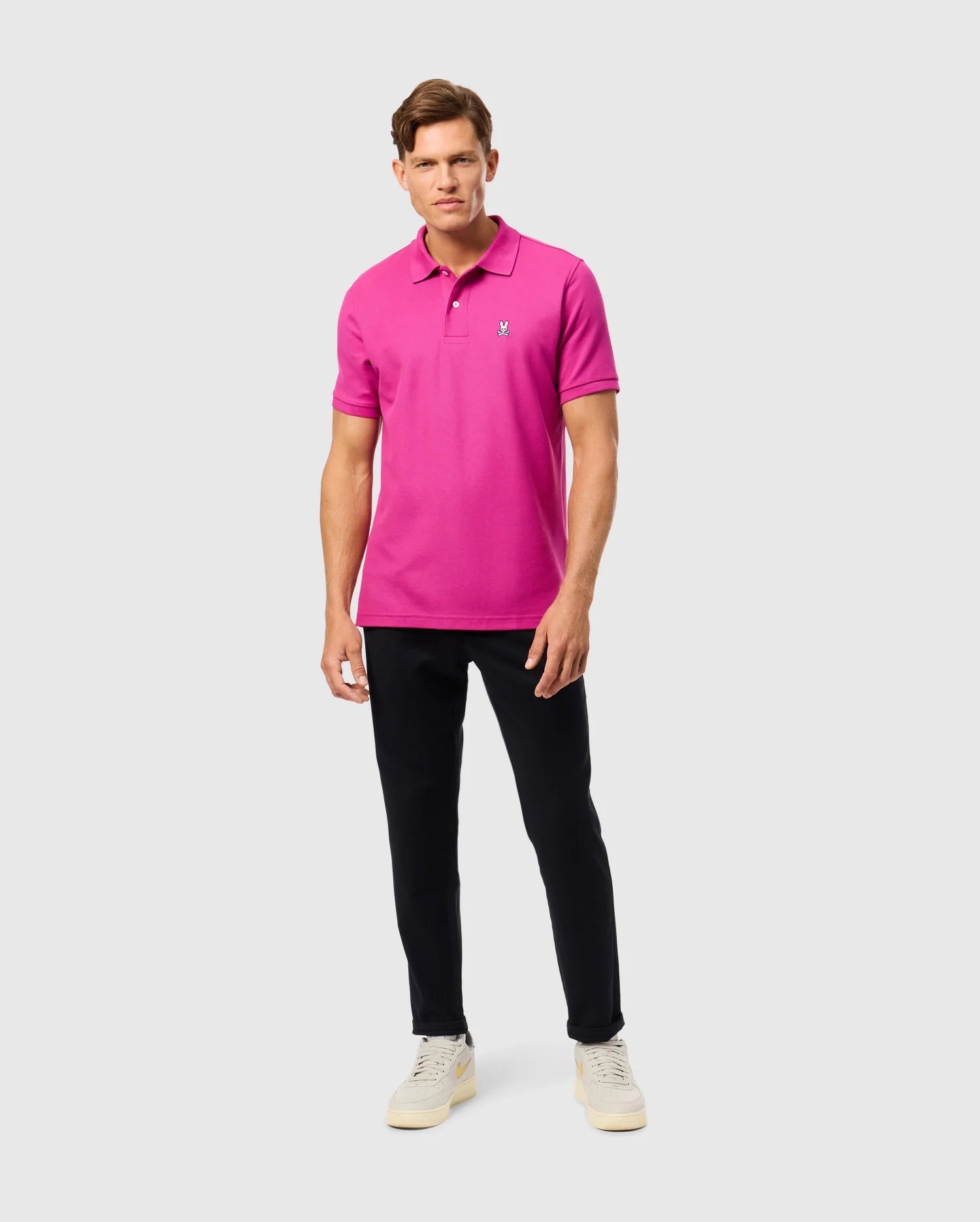 A person wearing the Psycho Bunny MENS CLASSIC PIQUE POLO SHIRT - B6K001D200 in a vibrant pink hue stands against a plain background. Paired with black pants and white sneakers, the 100% Pima cotton fabric exudes subtle elegance as they gaze ahead with a neutral expression.