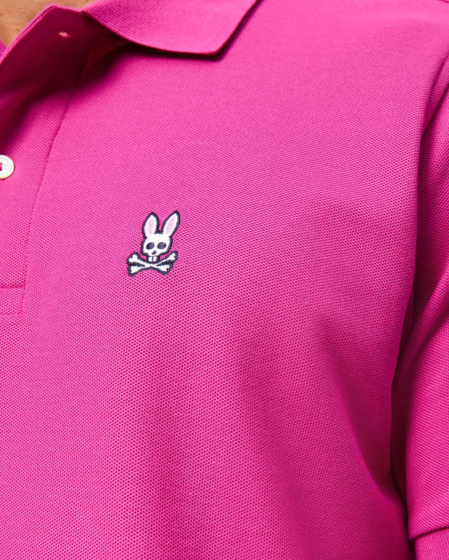 A close-up shows someone wearing the MENS CLASSIC PIQUE POLO SHIRT - B6K001D200 by Psycho Bunny. This classic pink shirt is crafted in Peru from 100% Pima cotton and features a textured fabric, white buttons, and a small logo of a cartoon rabbit's head over crossed bones stitched on the chest.