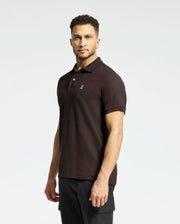 A man stands against a plain white background wearing a Psycho Bunny MENS CLASSIC PIQUE POLO SHIRT - B6K001D200 in dark brown, made from luxurious Pima cotton with a small embroidered logo on the chest, paired with black pants.