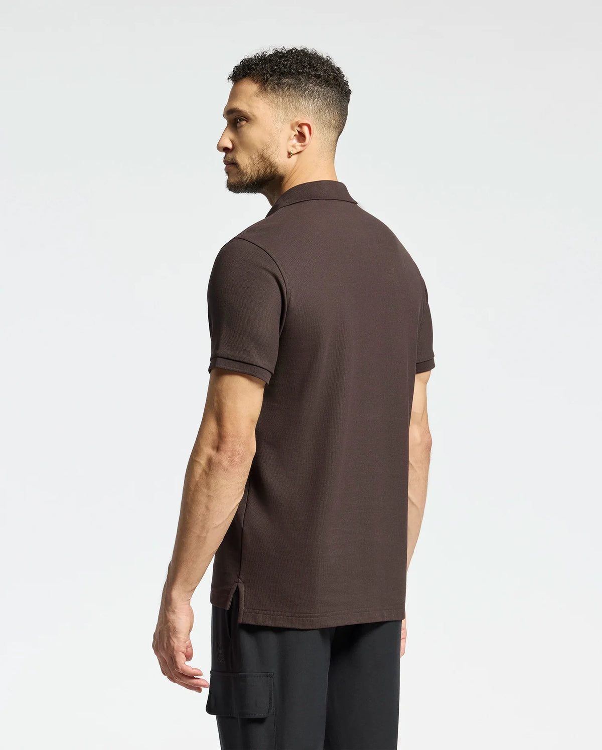 A man with short curly hair wears a Psycho Bunny MENS CLASSIC PIQUE POLO SHIRT - B6K001D200 and black pants. He stands to the side against a plain white background, showcasing timeless style and comfort.