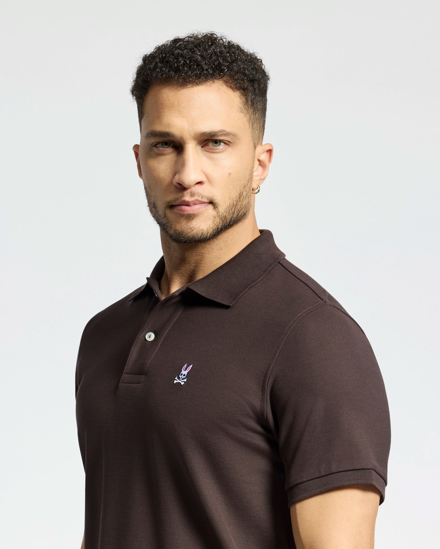 A man with short curly hair and a trimmed beard wears a Psycho Bunny MENS CLASSIC PIQUE POLO SHIRT - B6K001D200, featuring an embroidered logo. He has a neutral expression and a hoop earring, with the diamond-knit piqué fabric set against a plain, light-colored background.