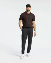 A man wears a black cap, Psycho Bunny's MENS CLASSIC PIQUE POLO SHIRT (B6K001D200) in dark, and black jogger pants. He stands against a plain white background, looking forward. White sneakers complete his casual outfit with hands resting by his sides.