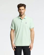 A man wears a Psycho Bunny Men's Classic Pique Polo (B6K001F200) in light green with an embroidered bunny logo on the chest, paired with black pants. He has short, styled hair and a neutral expression against a plain, light-colored background.