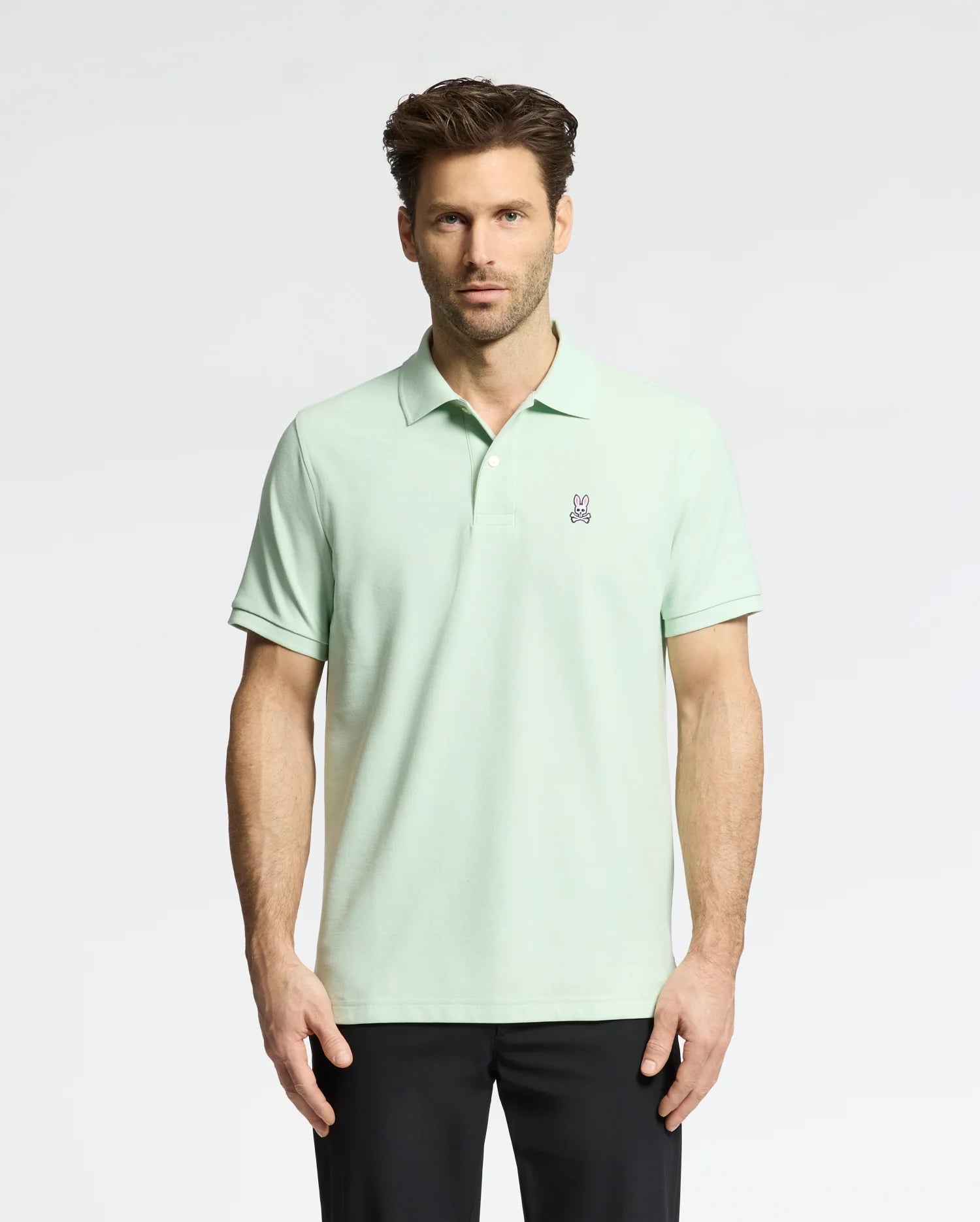 A man wears a Psycho Bunny Men's Classic Pique Polo (B6K001F200) in light green with an embroidered bunny logo on the chest, paired with black pants. He has short, styled hair and a neutral expression against a plain, light-colored background.