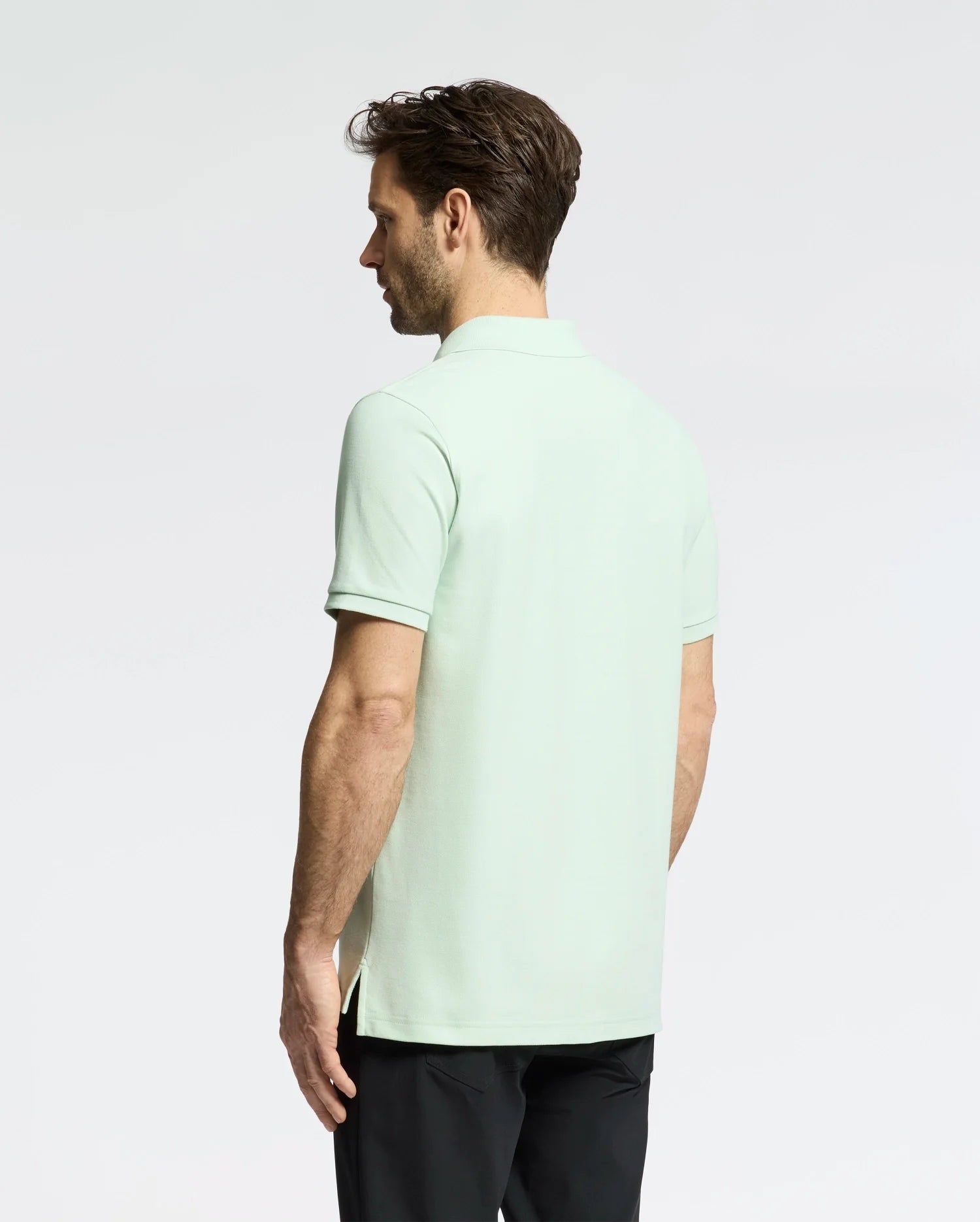 A dark-haired man stands with his back to the camera, wearing a light green Psycho Bunny Men's Classic Pique Polo (B6K001F200) with an embroidered bunny logo and black pants. The background is plain white.