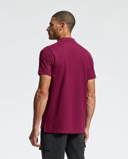 A man wearing a maroon Psycho Bunny Men's Classic Pique Polo Shirt (B6K001Z1PC) paired with black pants stands against a plain white backdrop. His head is slightly turned to the right, and his left arm hangs by his side, highlighting the elegance of the Pima cotton fabric adorned with an embroidered Bunny logo.