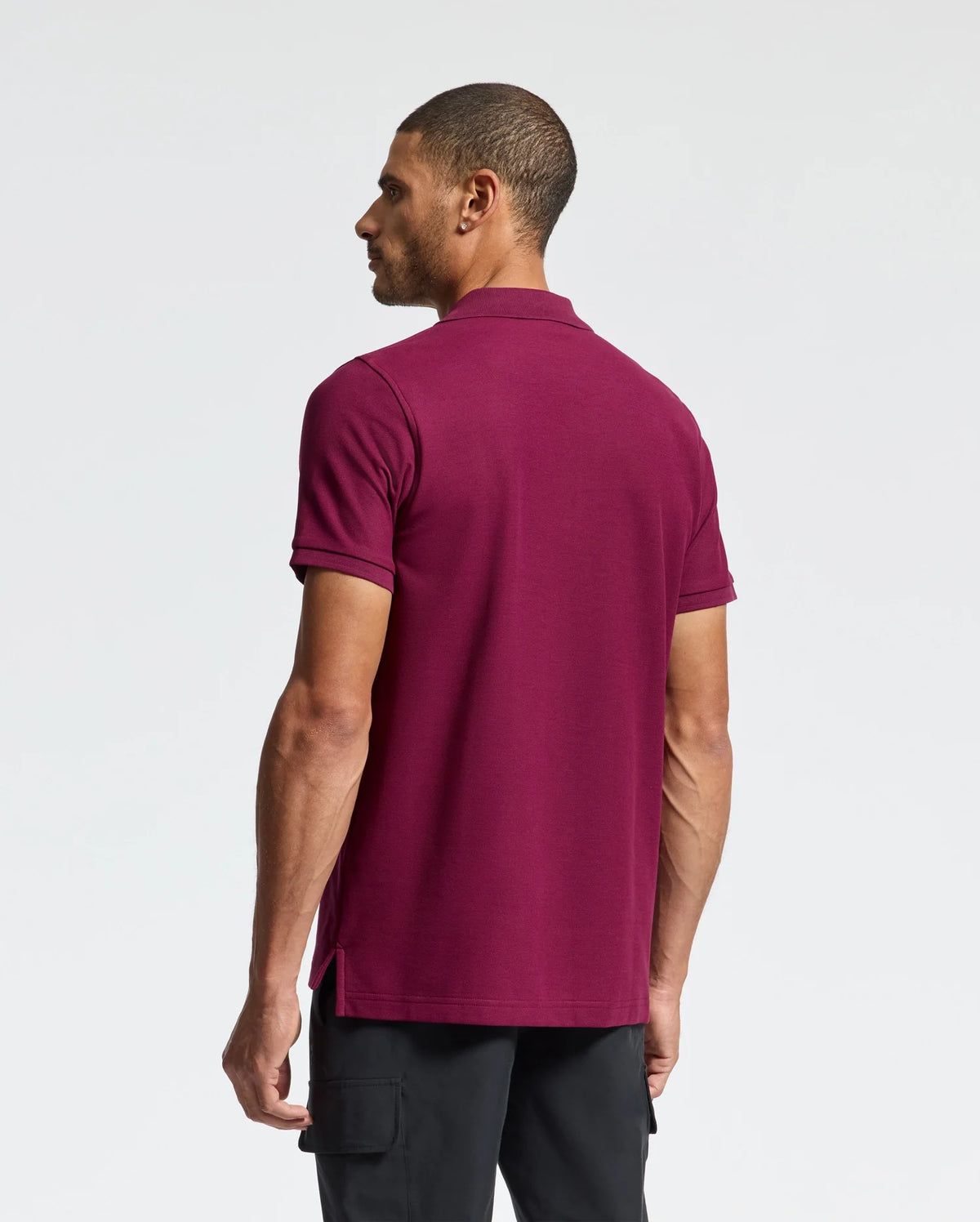 A man wearing a maroon Psycho Bunny Men's Classic Pique Polo Shirt (B6K001Z1PC) paired with black pants stands against a plain white backdrop. His head is slightly turned to the right, and his left arm hangs by his side, highlighting the elegance of the Pima cotton fabric adorned with an embroidered Bunny logo.