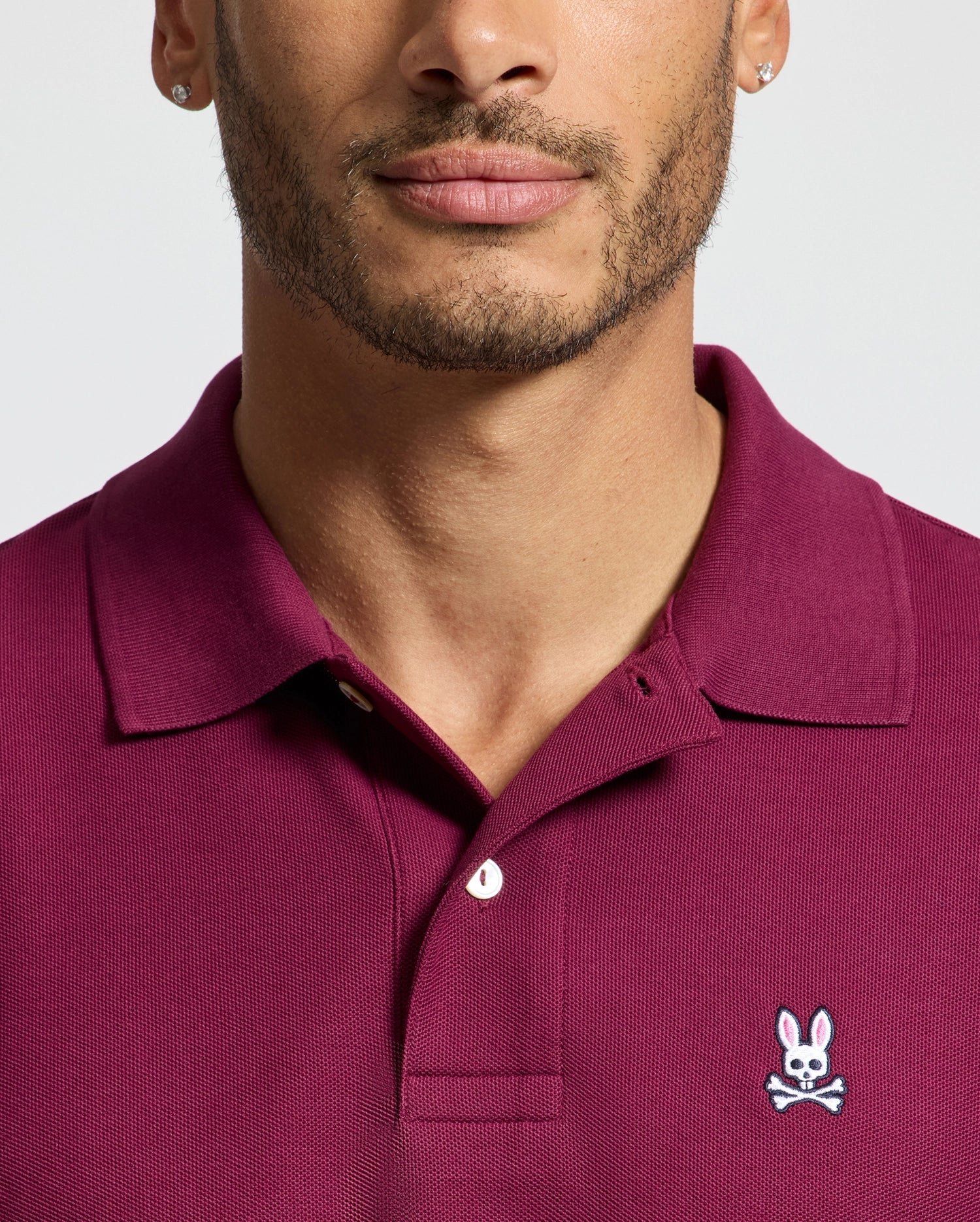 Someone is wearing a Psycho Bunny MENS CLASSIC PIQUE POLO SHIRT - B6K001Z1PC in burgundy, made from smooth Pima cotton with an embroidered Bunny logo on the chest, against a light gray background. The face is only partially visible.