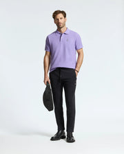 A man wearing a Psycho Bunny men's Classic Pique Polo (product code: B6K001Z1PC) in lavender, paired with black jogger pants and black boots, is holding a black bag. He poses against a plain white background.