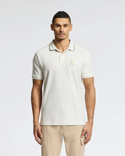 A man models the Psycho Bunny MENS ELDORADO PIQUE POLO - B6K105E200, made of Peruvian Pima cotton with a subtle chest logo. Standing against a white background, he pairs it with regular-fit light khaki pants, arms at his sides, gazing directly at the camera.