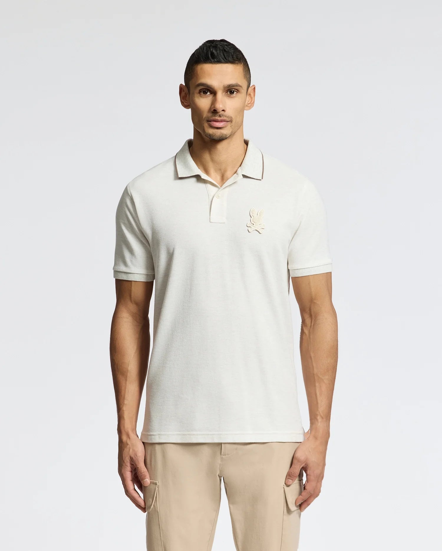 A man models the Psycho Bunny MENS ELDORADO PIQUE POLO - B6K105E200, made of Peruvian Pima cotton with a subtle chest logo. Standing against a white background, he pairs it with regular-fit light khaki pants, arms at his sides, gazing directly at the camera.