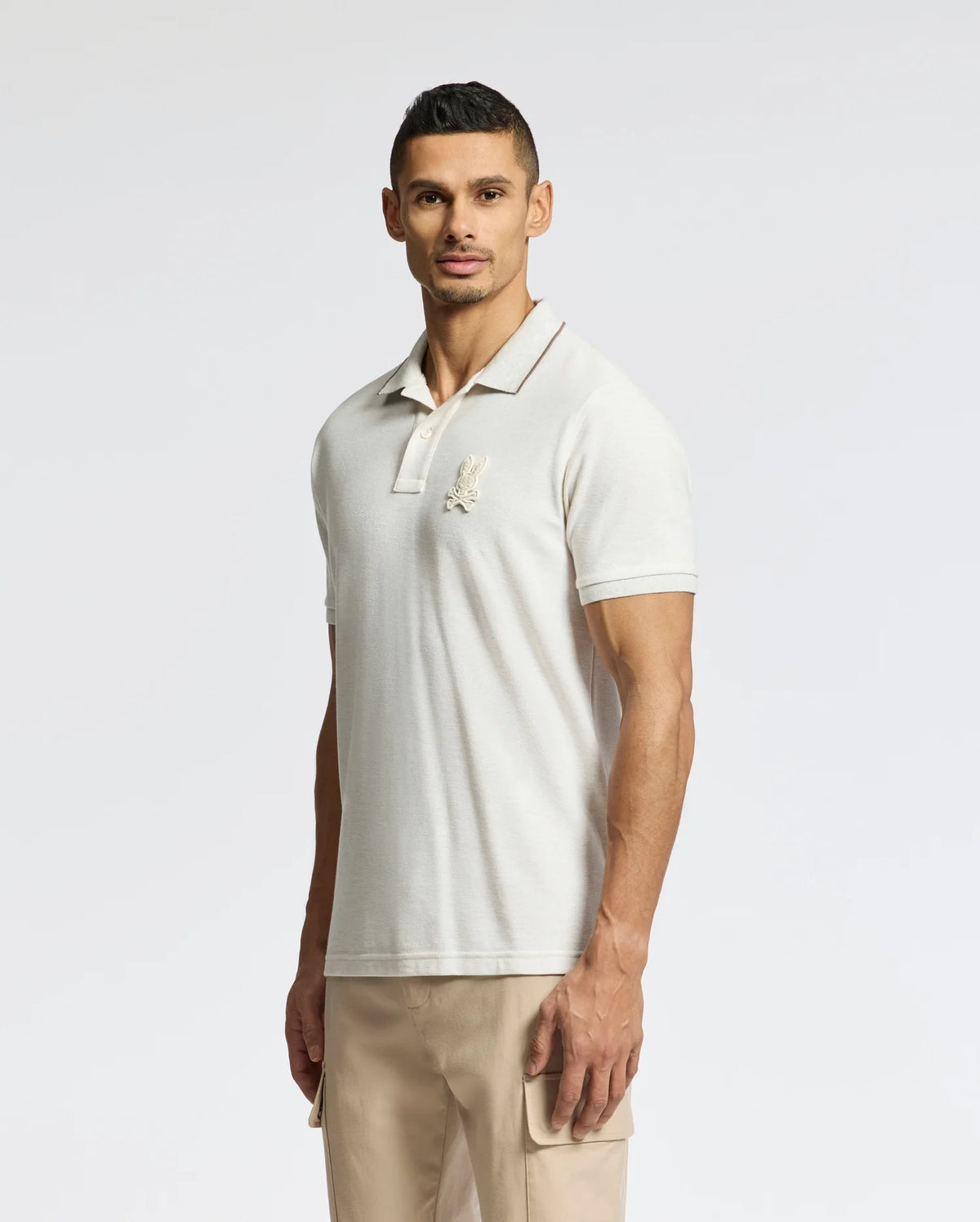 A man wears a Psycho Bunny MENS ELDORADO PIQUE POLO - B6K105E200 in light beige, made from luxurious Peruvian Pima cotton with a logo on the chest, paired with beige regular-fit pants. He stands against a white background, smiling slightly while facing the camera.