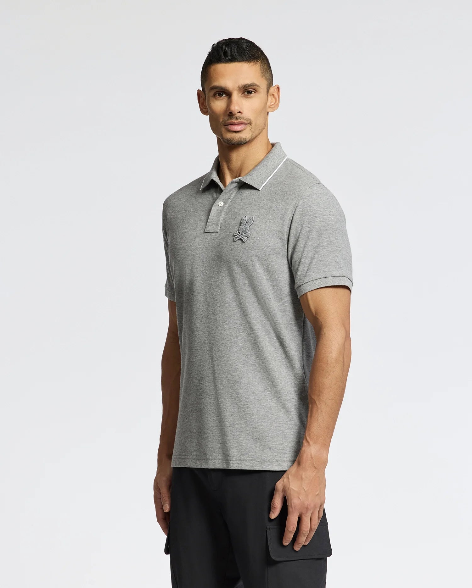 A man in a Psycho Bunny MENS ELDORADO PIQUE POLO - B6K105E200, made of gray Peruvian Pima cotton with an embroidered chest logo, stands against a plain background. He has short dark hair and wears black pants.