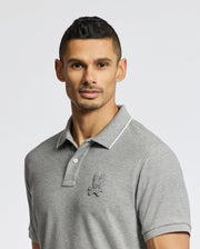A person with short dark hair and a neutral expression wears the Psycho Bunny MENS ELDORADO PIQUE POLO - B6K105E200, featuring gray Peruvian Pima cotton, a collar, buttons, and an embroidered chest design, against a plain backdrop.