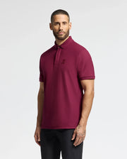 A man with short hair and a beard is wearing the luxurious MENS BRANSON PIQUE POLO by Psycho Bunny in burgundy, paired with black pants. He stands against a plain white background, looking directly at the camera with a neutral expression.