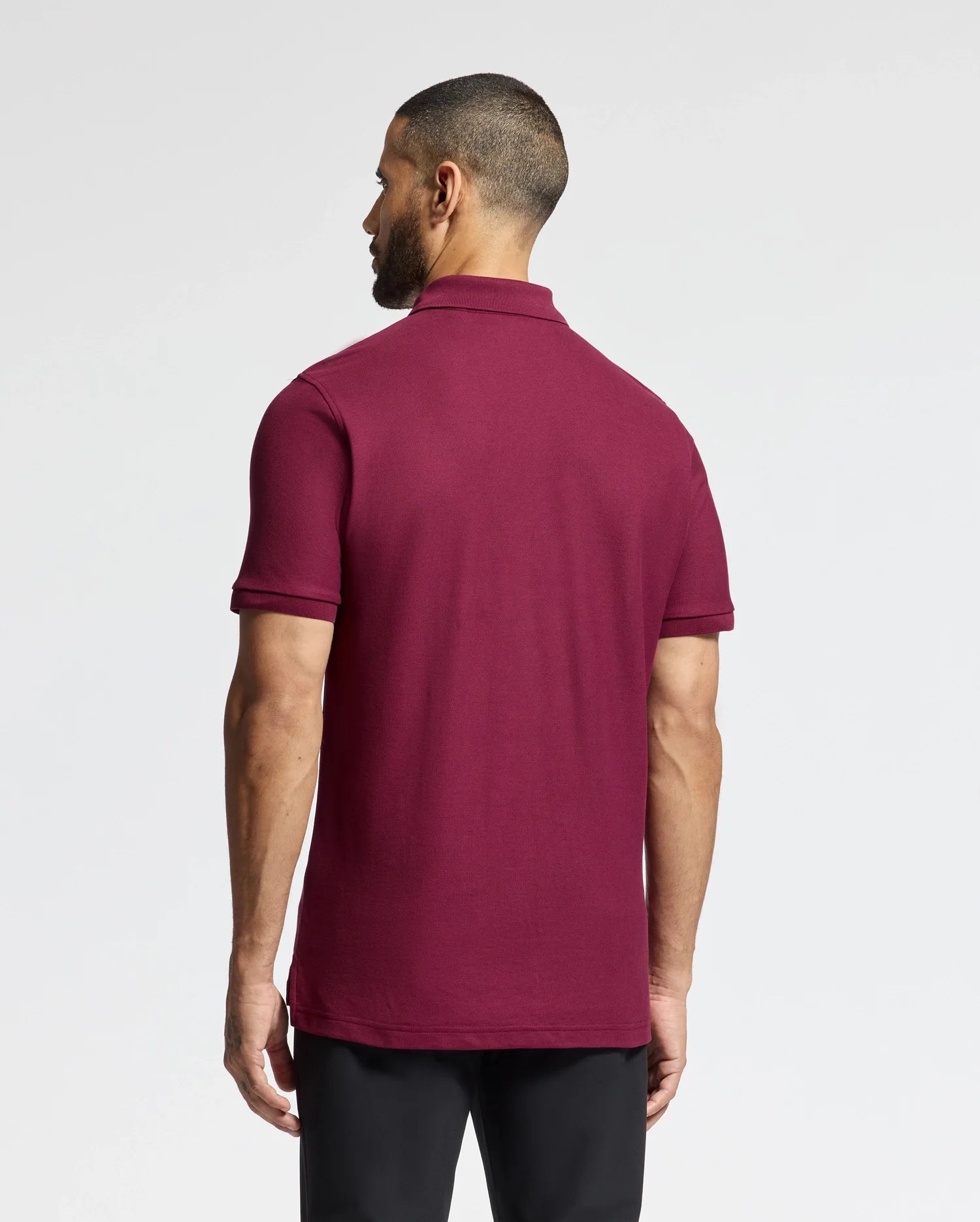 A man with short hair faces away from the camera, dressed in a luxurious Psycho Bunny MENS BRANSON PIQUE POLO - B6K108E200 in maroon, paired with black pants against a plain white background.