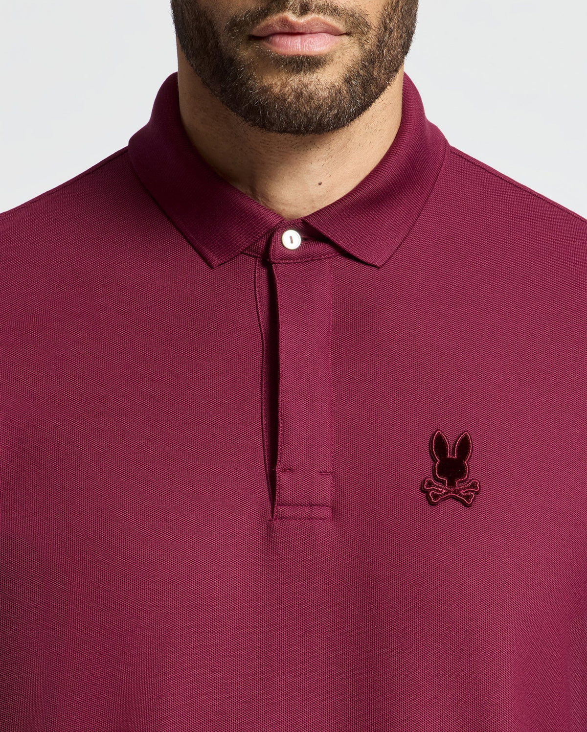 A person wearing a maroon Psycho Bunny Men's Branson Pique Polo (B6K108E200) with an embroidered rabbit head and crossed bones design on the left chest, and the shirt collar buttoned up.