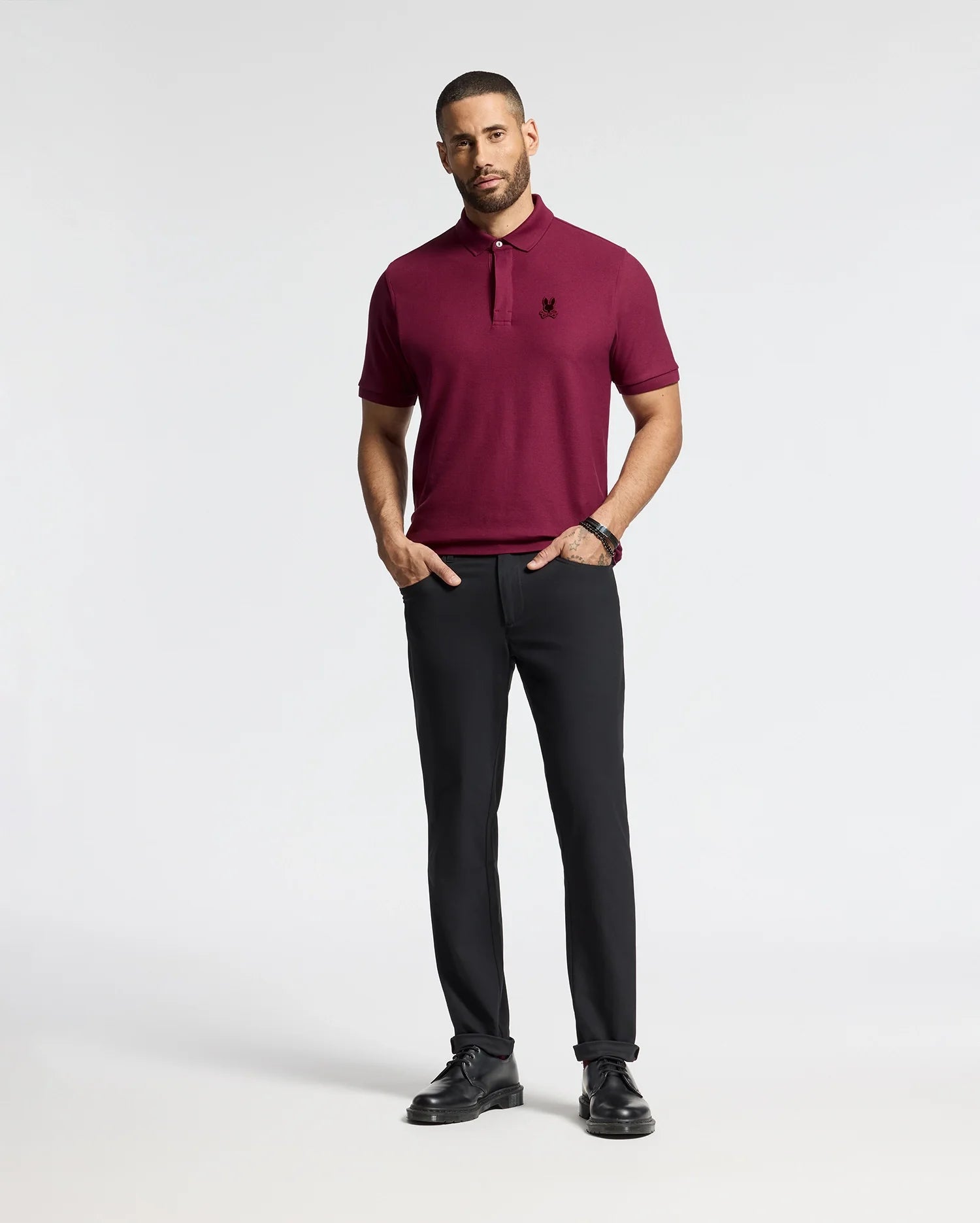 A man stands against a plain background wearing the luxurious Psycho Bunny Men's Branson Pique Polo in maroon, paired with black trousers. He has short hair and a beard, complemented by black shoes and a watch. His hands rest casually by his sides.