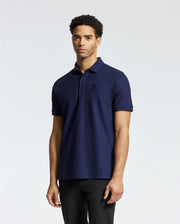A person wearing a Psycho Bunny MENS BRANSON PIQUE POLO - B6K108E200 in navy, featuring a Velvet Patch Logo on the left side, paired with black pants, stands against a plain light gray background.