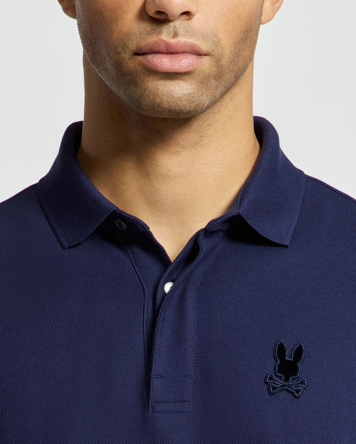 A person is wearing the Psycho Bunny MENS BRANSON PIQUE POLO - B6K108E200, crafted from premium navy blue Pima cotton and featuring an embroidered black bunny with crossbones logo on the chest. The image is cropped to highlight only the upper body.