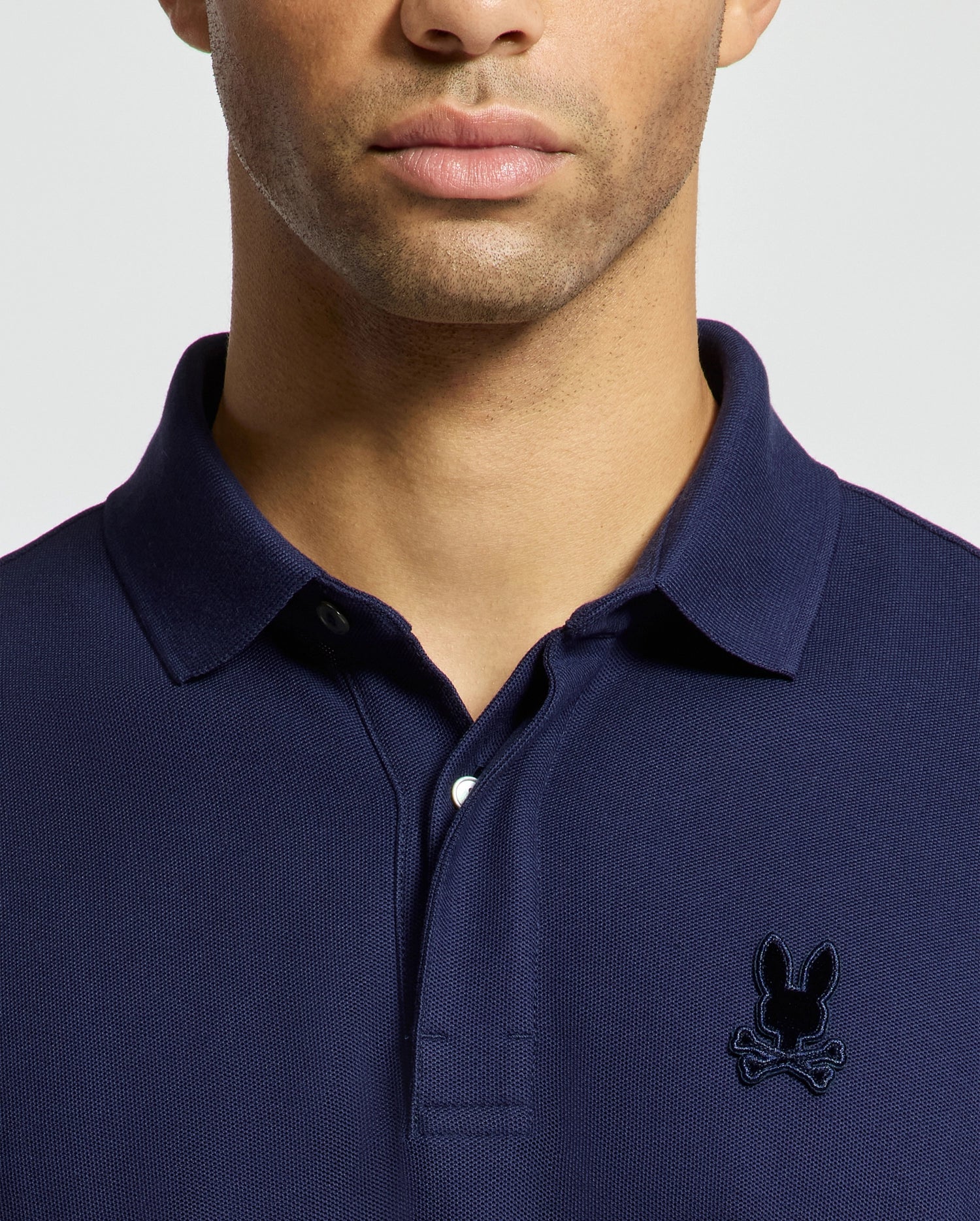A person is wearing the Psycho Bunny MENS BRANSON PIQUE POLO - B6K108E200, crafted from premium navy blue Pima cotton and featuring an embroidered black bunny with crossbones logo on the chest. The image is cropped to highlight only the upper body.