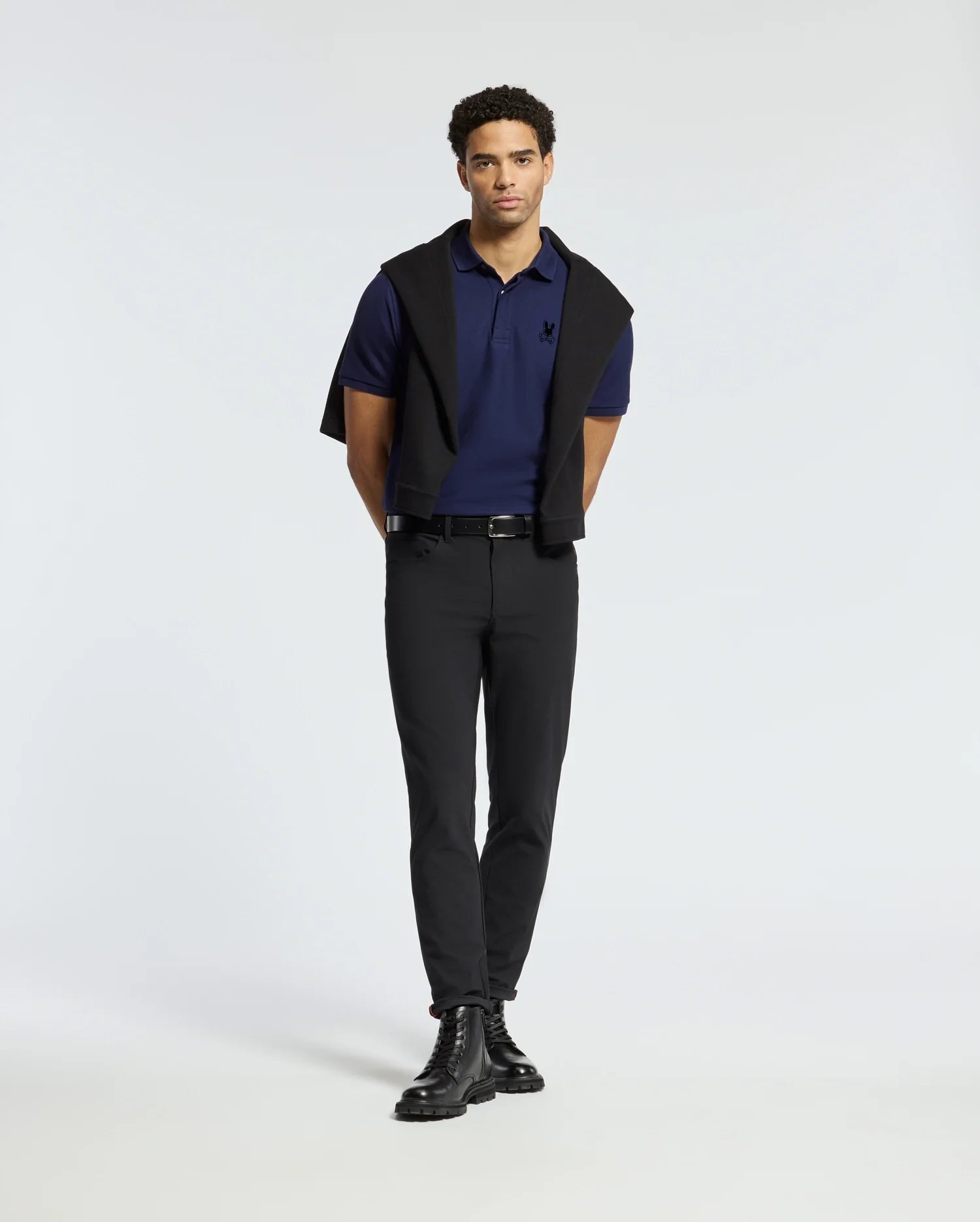 A man stands against a neutral background, showcasing the premium Psycho Bunny Men's Branson Pique Polo in navy, paired stylishly with black pants and boots. A black sweater is elegantly draped over his shoulders with the sleeves tied in front. His short curly hair and direct gaze enhance the sophisticated appearance.