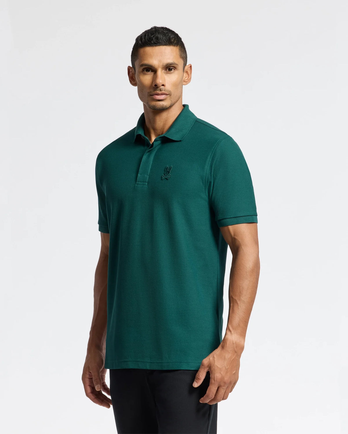 A person wearing a Psycho Bunny MENS BRANSON PIQUE POLO - B6K108E200 in teal, featuring a subtle logo on the chest, stands against a plain white background. The classic polo shirt is paired with dark pants, complementing their short, dark hair as they look directly at the camera.