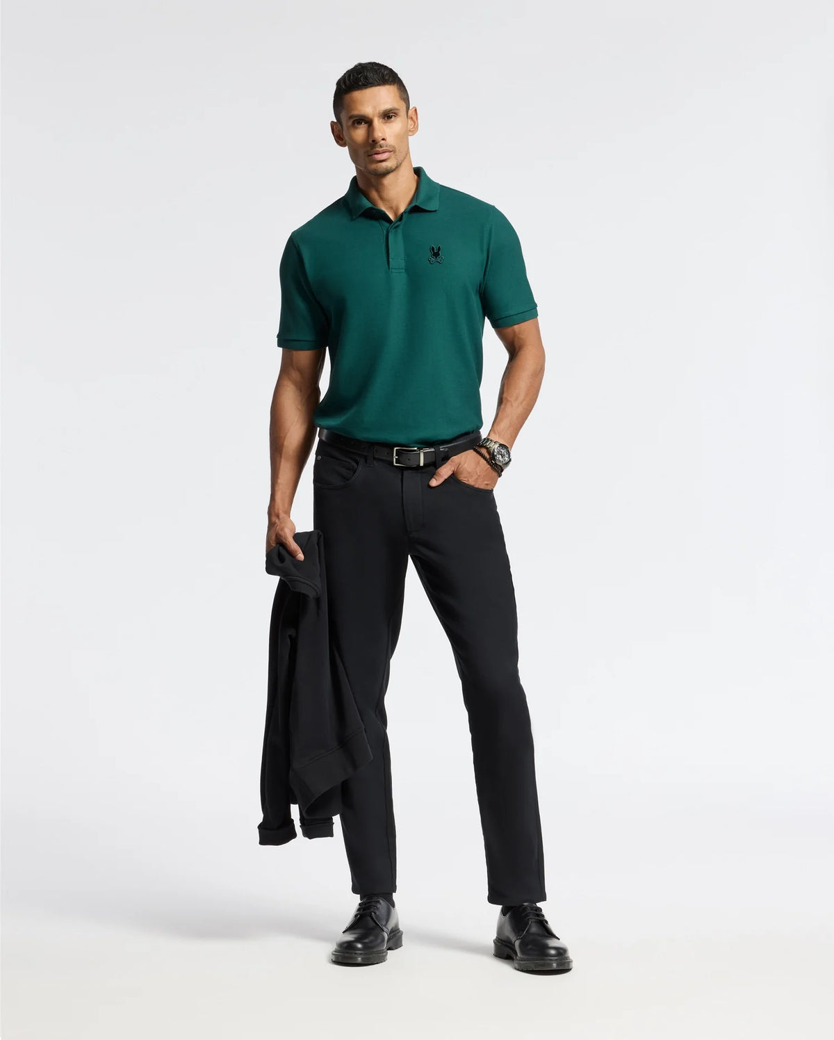 A man stands confidently against a white background, wearing a premium quality Psycho Bunny Men's Branson Pique Polo (B6K108E200) in green, paired with black pants and shoes. He casually holds a black jacket in his left hand while sporting a watch on his wrist.