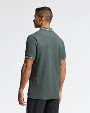 A man with short black hair is wearing the Psycho Bunny MENS PIERCE PIQUE POLO - B6K201E200 in dark green, which features white stripes on the sleeves and collar. The shirt is adorned with an embroidered Bunny logo, adding a touch of flair as he stands angled to the side, looking away against a plain white background.