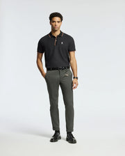 A man wearing a luxurious Psycho Bunny Men's Brandon Pique Polo in black, olive green trousers, and black shoes stands against a plain background.