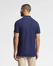 A man with short hair and a beard stands facing away, showcasing the luxurious Psycho Bunny MENS BRANDON PIQUE POLO - B6K280E200. The navy blue shirt features light blue trimming on the sleeves and collar, paired stylishly with beige pants. The plain white background highlights the premium quality of his attire.