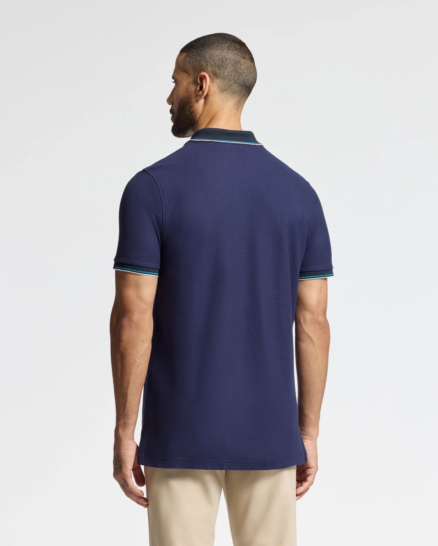 A man with short hair and a beard stands facing away, showcasing the luxurious Psycho Bunny MENS BRANDON PIQUE POLO - B6K280E200. The navy blue shirt features light blue trimming on the sleeves and collar, paired stylishly with beige pants. The plain white background highlights the premium quality of his attire.